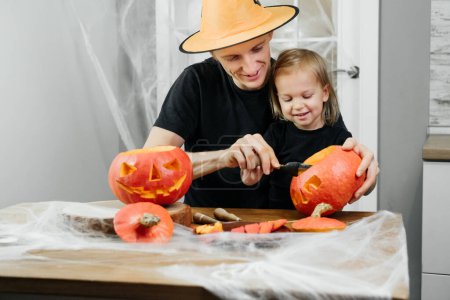 Foto de Los padres con chld están tallando Jack o linterna de calabaza para Halloween en casa. Padre en sombrero preparándose para la selebración con su hija. - Imagen libre de derechos