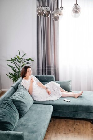 Photo for Woman drinking mug of coffee in cozy home atmosphere in the morning. Real middle age plus size female in white dress relaxing on the sofa, modern house. Sunny day with cup of tea, laying on couch - Royalty Free Image