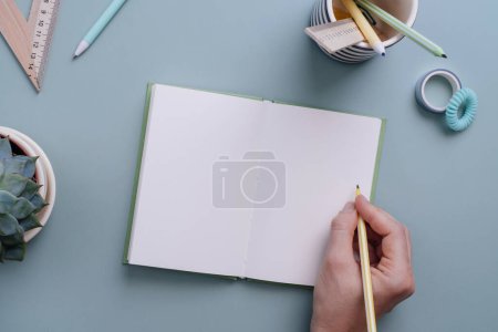 Foto de Mano de mujer lista para escribir en el cuaderno vacío sobre el fondo azul pastel. Lugar para el texto. Mesa de trabajo para trabajo creativo, estudio o planificación - Imagen libre de derechos