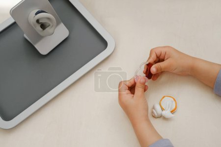 Photo for Child holding hearing aids in medical clinic, ear model on the background. Kid with hearing loss tring listening device. Closeup of hands. Top view - Royalty Free Image
