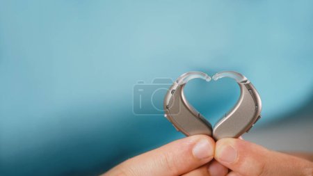 Prothèses auditives dans les mains faisant forme de coeur sur fond bleu. Gros plan de l'appareil d'écoute pour les personnes ayant un trouble auditif, un dysfonctionnement. Technologie qui donne un meilleur sens du son, speach