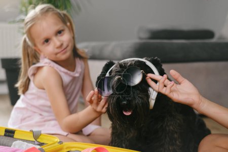 Téléchargez les photos : Un enfant joue avec un chien dans un casque. Fille S'amuser avec l'animal tout en se préparant pour un voyage. Chien en lunettes de soleil écoutant de la musique, partant en vacances - en image libre de droit