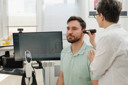 Man has appointment at clinic, checking hearing. Doctor audiologist consulting at hearing rehabilitation clinic to select digital device for middle age man. Doctor consulting patient