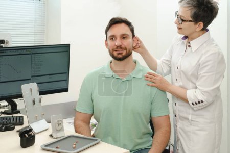 Photo for Man trying modern compact hearing aids at clinic. Audiologist consulting at hearing rehabilitation clinic to select digital device for middle age man. Doctor consulting patient - Royalty Free Image