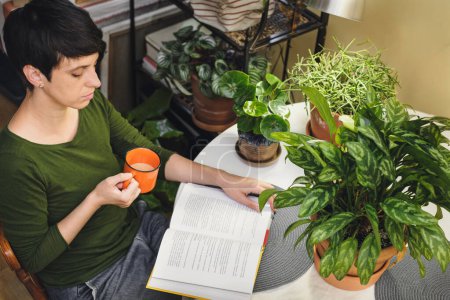Photo for Woman at her reading place near houseplants. Green relax zone in the room with opened book and potted flowers. Female Enjoying peace and having rest at eco designed cozy house. - Royalty Free Image