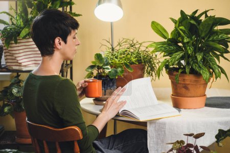 Photo for Woman at her reading place near houseplants. Green relax zone in the room with opened book and potted flowers. Female Enjoying peace and having rest at eco designed cozy house. - Royalty Free Image