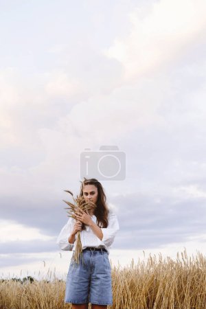 Téléchargez les photos : Femme marchant sur le champ d'or de blé tenant tas de seigle. Femme à la mode Toucher l'oreille de blé à l'extérieur le soir. Espace de copie - en image libre de droit