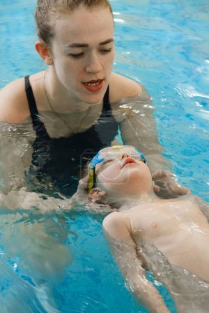 Téléchargez les photos : Enfant en bas âge apprenant à nager dans la piscine intérieure avec professeur. Flottant dans l'eau, équilibrage et activité physique générale pour les enfants, développement précoce. Garçon formé pour botter les jambes et flotter - en image libre de droit