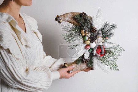 Téléchargez les photos : Gros plan femme tenant une couronne de Noël en forme de lune avec elfe. La base de la couronne est branches d'épinette, pas de déchets décoration festive dans les mains des femmes. Brunettes vertes avec des bobbles, gros plan sur blanc - en image libre de droit