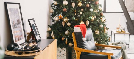 Foto de Acogedor sillón cerca del árbol de Navidad en habitación soleada con grandes ventanales. Sombrero de Santa por la mañana. A nadie. espacio de copia. Banner de saludo - Imagen libre de derechos