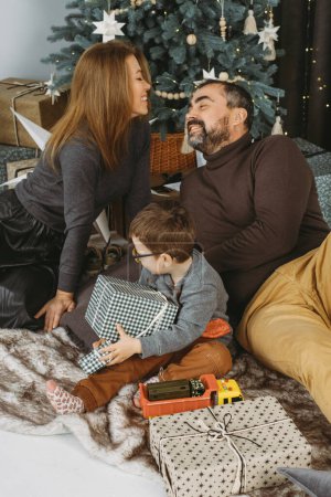 Téléchargez les photos : Famille avec enfant jouant près de l'arbre de Noël. Enfant déballer des cadeaux, les parents d'âge moyen profiter de l'humeur de Noël. Célébration à la maison - en image libre de droit