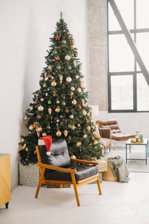 Foto de Acogedor sillón cerca del árbol de Navidad en habitación soleada con grandes ventanales. Sombrero de Santa por la mañana. A nadie. espacio de copia. Tarjeta postal de felicitación - Imagen libre de derechos