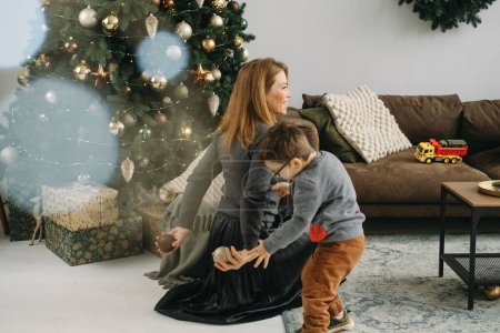 Téléchargez les photos : Mère avec un enfant jouant près de l'arbre de Noël. Enfant et femme d'âge moyen profiter de l'humeur de Noël. Fête à la maison. Bokeh. Espace de copie - en image libre de droit