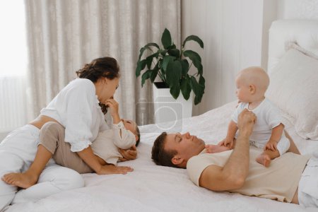 Photo for Family with two kids on the bed having fun. Mather, father baby and toddler sons are happy in the morning in bedroom. Children playing with parents at home. Real life and happiness - Royalty Free Image