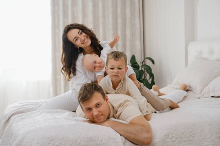 Photo for Family with two kids on the bed having fun. Mather, father baby and toddler sons are happy in the morning in bedroom. Children playing on fathers back. Real life and happiness - Royalty Free Image