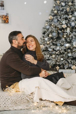 Photo for Happy couple celebrating romantic Christmas eve at home. Christmas interior decoration for family party. Copy space, greeting card. Middle-aged man and woman sitting on cozy bed with knitted blanket - Royalty Free Image