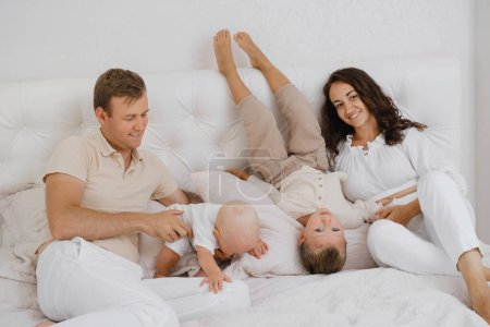 Téléchargez les photos : Famille avec deux enfants sur le lit s'amuser. Mather, père bébé et tout-petit fils sont heureux le matin dans la chambre. Les enfants jouent avec leurs parents à la maison. Vie réelle et bonheur - en image libre de droit