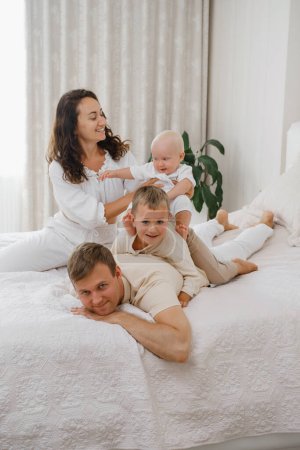 Téléchargez les photos : Famille avec deux enfants sur le lit s'amuser. Mather, père bébé et tout-petit fils sont heureux le matin dans la chambre. Les enfants jouent avec leurs parents à la maison. Vie réelle et bonheur - en image libre de droit