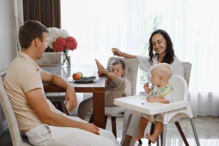 Téléchargez les photos : Famille avec enfants mangeant à table à la maison. Mère nourrissant ses frères et sœurs avec des collations saines dans une atmosphère détendue et amusante. Aliments sains pour enfants - en image libre de droit