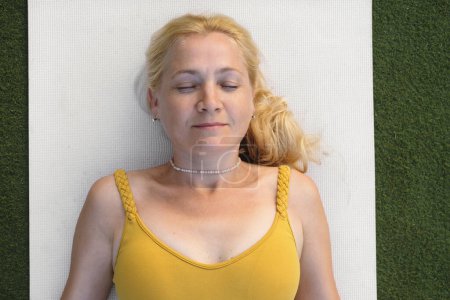 Téléchargez les photos : Femme relaxante dans la pose de shavasana après la pratique du yoga. Soins de santé préventifs pour adultes. Pleine conscience. Femme dans la cinquantaine allongée sur un tapis. Vue du dessus - en image libre de droit
