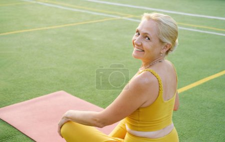 Téléchargez les photos : Femme caucasienne mature méditant en plein air sur le coucher du soleil sur le terrain de sport. Femme âgée regardant la caméra tout en étant assis dans la pose de lotus, faire du yoga dans le parc - en image libre de droit