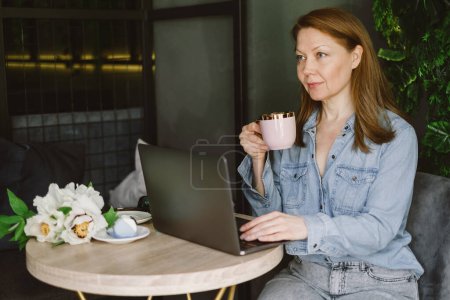 Foto de Mujer trabajando en el portátil en la cafetería. Freelancer de mediana edad con taza de café escribiendo y leyendo. Loft lugar de trabajo con plantas verdes - Imagen libre de derechos