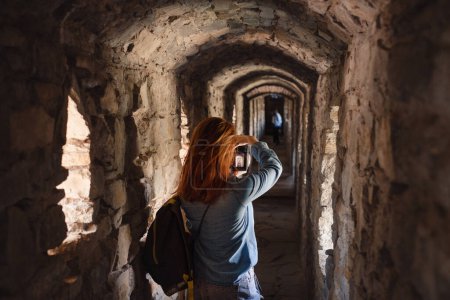 Foto de Mujer caminando por el viejo túnel, mirando por la ventana y tomando fotos. Castillo de visita femenina. Viajar en un edificio antiguo - Imagen libre de derechos