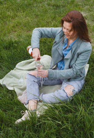 Foto de La mujer descorcha la botella de vino rosa en el picnic. Hermosa mujer madura servir vino en vidrio al aire libre en la hierba. Mujer de 50 años en el parque - Imagen libre de derechos