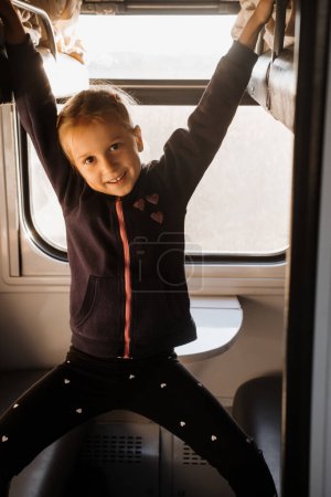 Foto de Feliz niño cabalgando en tren en la puesta del sol, la luz del sol brillante, viajes atmosféricos en tren con los niños. Pasajero joven jugando en el sofá de la carreta resbaladiza. Largo viaje familiar - Imagen libre de derechos