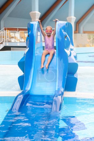 Téléchargez les photos : Enfant glissant dans l'eau dans un parc aquatique. Enfant s'amusant et divertissant dans la piscine intérieure. Jeune fille faisant des éclaboussures - en image libre de droit
