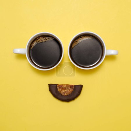 Téléchargez les photos : Café créatif heureux emoji. Sourire de deux tasses d'expresso noir et dessert cru sain en bouche. Visage souriant avec les yeux sur fond jaune. Vue de dessus. Une nourriture qui apporte des émotions positives - en image libre de droit