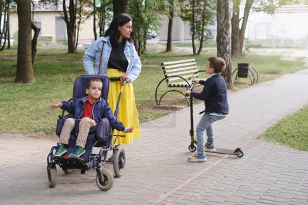 Foto de Familia con niño que tiene parálisis cerebral, usuario de silla de ruedas que camina al aire libre. Integración y accesibilidad de las personas con discapacidad, inclusión. Hermano con trastorno físico - Imagen libre de derechos