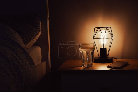 Téléchargez les photos : Lampe près du lit. Lumière tamisée dans la pièce. Éclairage de lanterne. Bonne nuit de sommeil routine avec gadgets éteints et de l'eau pour l'hydratation - en image libre de droit