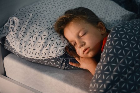 Téléchargez les photos : Enfant dormant au lit tard dans la nuit. Mignon oreiller fille tout-petit recouvert d'une couverture chaude. Le gamin se repose la nuit. Lumière sur la table de nuit - en image libre de droit