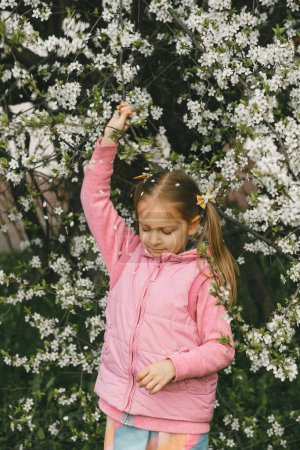 Photo for Nice little child in blooming garden exploring spring nature. Shaking tree making petals falling on her head. Fun in the garden. - Royalty Free Image