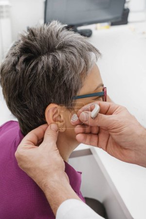 Téléchargez les photos : Otolaryngologue mettant des aides auditives miniatures modernes pour femme d'âge moyen au centre médical moderne. Audiologiste aide à évaluer les troubles de l'audition Aides auditives invisibles petites et discrètes dans les mains - en image libre de droit