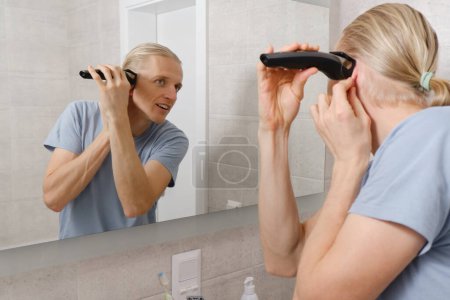 Téléchargez les photos : Homme rasant moustaches cheveux par lui-même, coupe de cheveux à la maison devant le miroir dans le bain. Homme avec une longue coupe de cheveux à l'aide d'une tondeuse à raser. Rasoir électrique pour l'hygiène personnelle en quarantaine - en image libre de droit