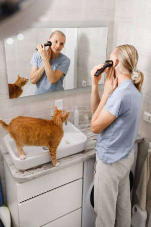 Foto de Hombre afeitando bigotes pelo por sí mismo, auto corte de pelo en casa delante del espejo en el baño con el gato. Trimmer máquina de afeitar maleusing. maquinilla de afeitar eléctrica para la higiene personal en cuarentena - Imagen libre de derechos