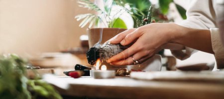 Photo for Woman hands burning white sage, palo santo before ritual on the table with candles and green plants. Smoke of smudging treats pain and stress, clear negative energy - Royalty Free Image