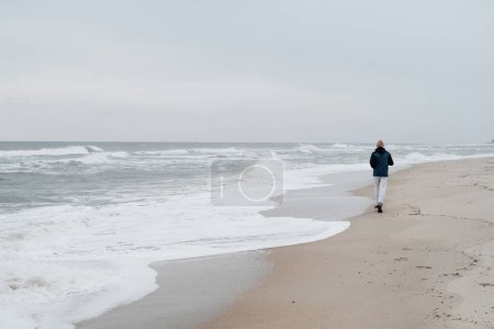 Téléchargez les photos : Homme d'âge moyen courant sur la plage en automne. Repos hors saison sur la mer, mode de vie sain. Athlète coureur en veste chaude à l'automne près de l'eau. Entraînement de coureur masculin en plein air jogging dans la nature - en image libre de droit