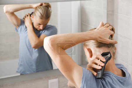 Téléchargez les photos : Hygiène personnelle, homme caucasien couper ses propres cheveux dans la salle de bain avec rasoir électrique sans fil devant le miroir - en image libre de droit