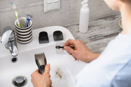 Photo for Man cleaning electronic shaving razor in sink, male self care and personal hygiene Top view. Hair in sink with cat. Long blonde hair, machine in hands - Royalty Free Image