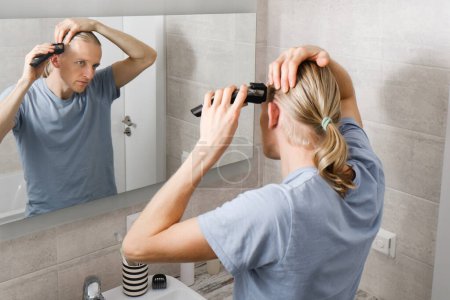 Foto de Higiene personal, hombre caucásico cortando su propio cabello en el baño con la máquina de afeitar eléctrica inalámbrica en frente del espejo - Imagen libre de derechos