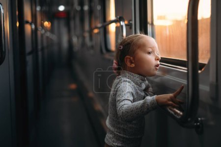 Photo for Toddler child looking though train window on sunset, bright sunlight, atmospheric travel by railway with kids. Girl happy exploring the way in evening. Exciting family trip - Royalty Free Image
