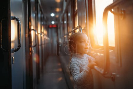 Foto de Niño pequeño que mira a través de la ventana del tren en la puesta del sol, la luz del sol brillante, los viajes atmosféricos en tren con los niños. Chica feliz explorando el camino en la noche. Emocionante viaje familiar - Imagen libre de derechos