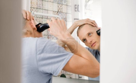 Foto de Higiene personal, hombre caucásico cortando su propio cabello en el baño con la máquina de afeitar eléctrica inalámbrica en frente del espejo. Vista desde la puerta. Copiar espacio - Imagen libre de derechos