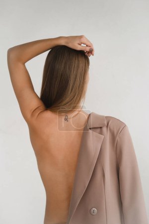 Photo for Pretty cheeky Caucasian woman tacking off business jacket. Sensual fashion style body of decisive strong female in minimalistic room. Naked back, shoulder covered by jacket - Royalty Free Image
