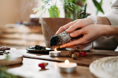Frauenhände verbrennen weißen Salbei, palo santo vor dem Ritual auf dem Tisch mit Kerzen und grünen Pflanzen. Rauchschwaden behandeln Schmerz und Stress, klare negative Energie