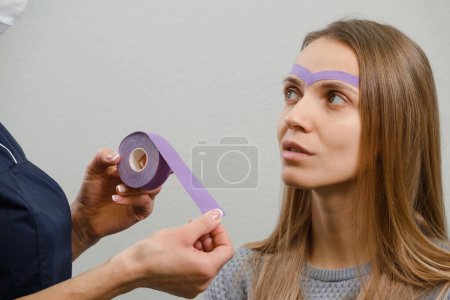 Foto de Cosmética femenina haciendo la cinta kinesio cosmética de la cara. Rayas púrpuras en la frente contra las arrugas y el envejecimiento. Medicina alternativa para la belleza en el hogar - Imagen libre de derechos