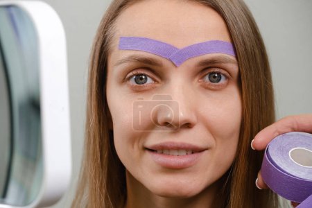 Photo for Female cosmetician doing the cosmetic kinesio taping of the face. Purple stripes on forehead against wrinkles and aging - Royalty Free Image